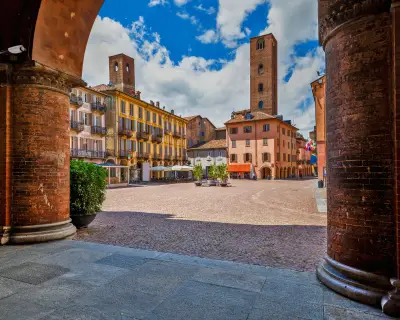View of cobblestone town square among old houses and medieval towers under beautiful sky in Alba, Piedmont, Northern Italy.