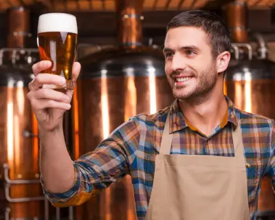 Happy brewer. Happy young male brewer in apron holding glass with beer and looking at it with smile while standing in front of metal containers