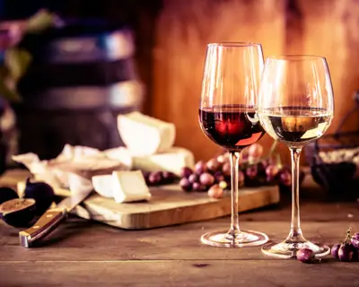 Cheese platter with fresh grapes and glasses of red and white wine on a rustic wooden table in front of a blazing fire in a tavern or winery