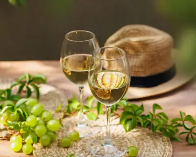 Two glasses of white wine with green grapes and straw hat on wooden patio table in natural light..